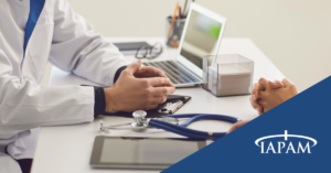 Clinical consultation scene with a doctor and patient at a desk, with a laptop, stethoscope, and tablet present, and IAPAM branding in the corner.
