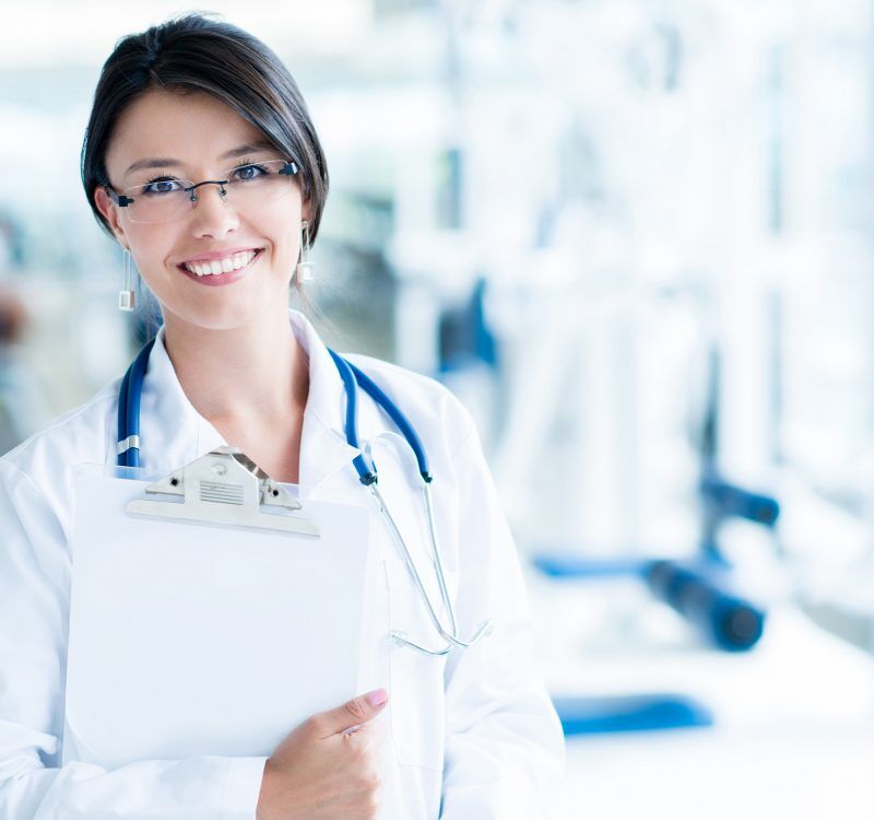 Female doctor in a white coat holding a clipboard, smiling in a bright clinical hallway with a stethoscope around her neck.