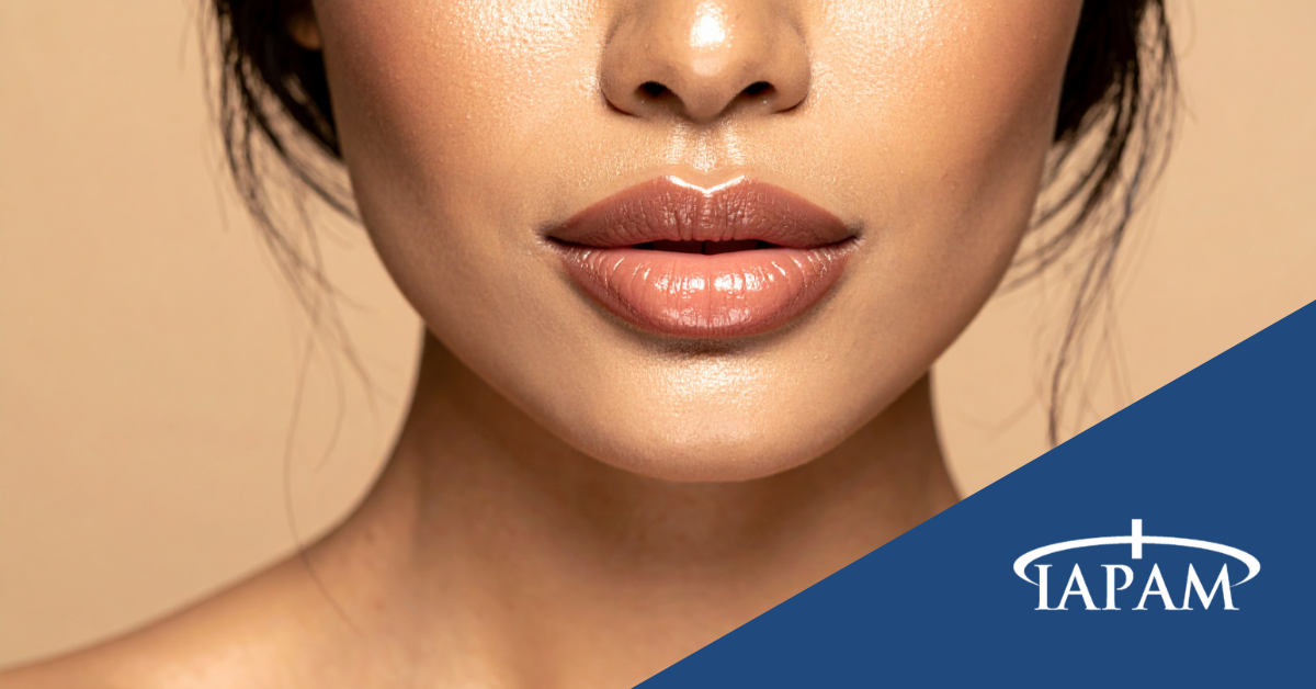 Close-up of a woman's glossy peach lips and chin against a beige background, with a blue diagonal banner showing the TAPAM logo in the bottom-right corner.