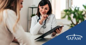 Doctor in a white coat with a stethoscope consults with a patient at a desk, examining information on a tablet, TAPAM branding in the blue corner.